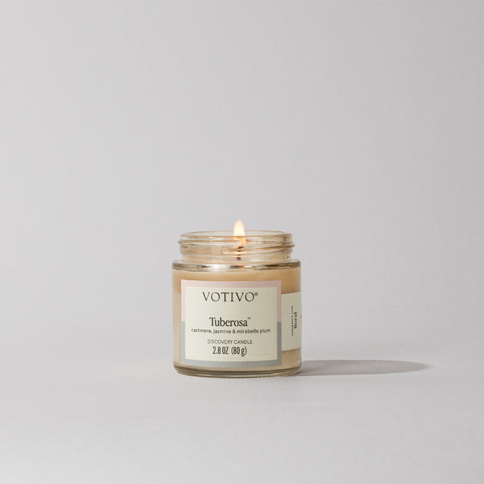 A small Tuberosa Discovery Candle by Votivo with a minimalist label sits lit on a light gray surface. This soy blend candle fills the air with an elegant tuberose fragrance.