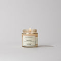A small Tuberosa Discovery Candle by Votivo with a minimalist label sits lit on a light gray surface. This soy blend candle fills the air with an elegant tuberose fragrance.