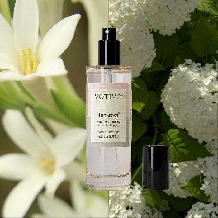 A Votivo Tuberosa Room & Linen Spray bottle with a black cap is displayed against white tuberose flowers on the left and white hydrangea blooms on the right, emphasizing its fresh scent.