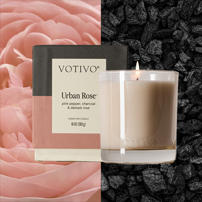 A Votivo Urban Rose 10 oz. Signature Candle and its box are shown, highlighting notes of pink pepper, charcoal, and damask rose. Pink rose petals and black charcoal pieces decorate the background. Made with a soy blend wax.