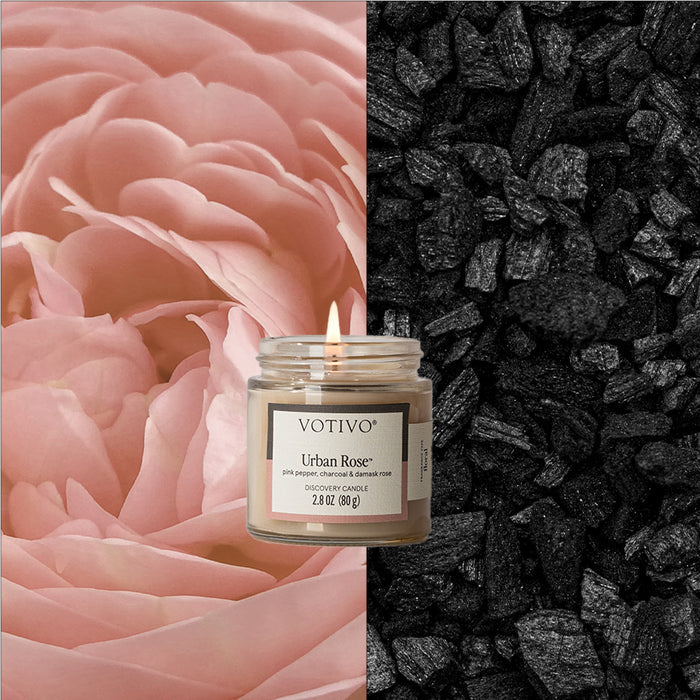 A lit Votivo Urban Rose Discovery Candle, made from soy blend wax, sits between a soft pink rose close-up on the left and a pile of black charcoal pieces on the right.