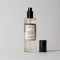 A clear bottle of Votivo Urban Rose Room & Linen Spray with a black nozzle and cap sits on a light gray background. The label notes charcoal rose, pink pepper, and damask rose in this distinctive urban fragrance.