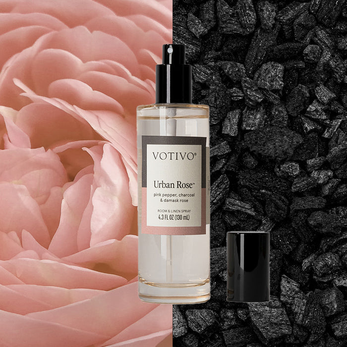 A clear Votivo Urban Rose Room & Linen Spray bottle, cap off, sits between a pink rose and dark charcoal pieces, capturing a unique charcoal rose vibe.