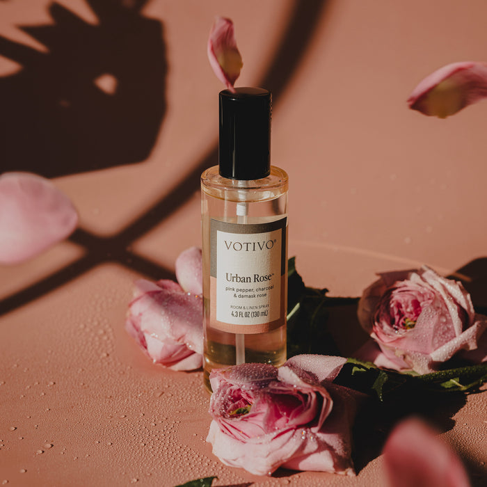 A bottle of Votivo Urban Rose Room & Linen Spray rests on a pink surface amid roses and petals, as gentle sunlight casts floral shadows across the scene.