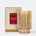 The Votivo Velvet Rouge Holiday Candle in a ribbed glass jar sits next to its beige and gold striped box, both with red labels detailing notes of cinnamon, red berries, and brown sugar. Made from premium soy blend wax.