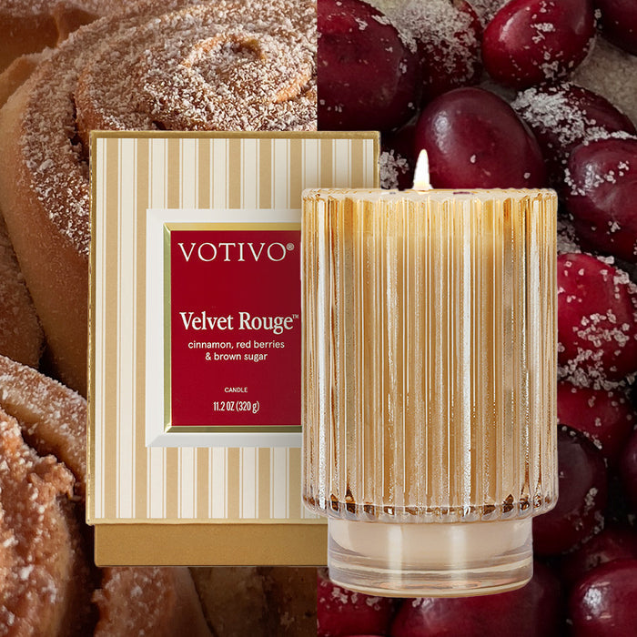 The Votivo Velvet Rouge Holiday Candle, made from soy blend wax, comes in a gold-ribbed glass jar with a striped box, set amid sugared cinnamon rolls and red berries.
