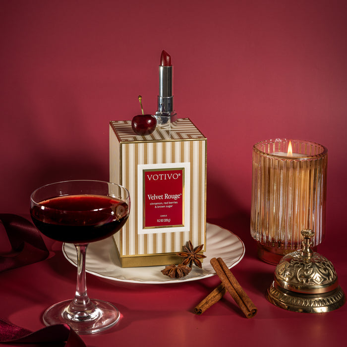 An artfully arranged display features the Votivo Velvet Rouge Holiday Candle—crafted from a luxurious soy blend wax—burning beside its striped box accented by a cherry-topped lipstick. The scene is completed with a glass of red wine, cinnamon sticks, star anise, and a festive bell, all presented on a white plate against a sumptuous red backdrop.