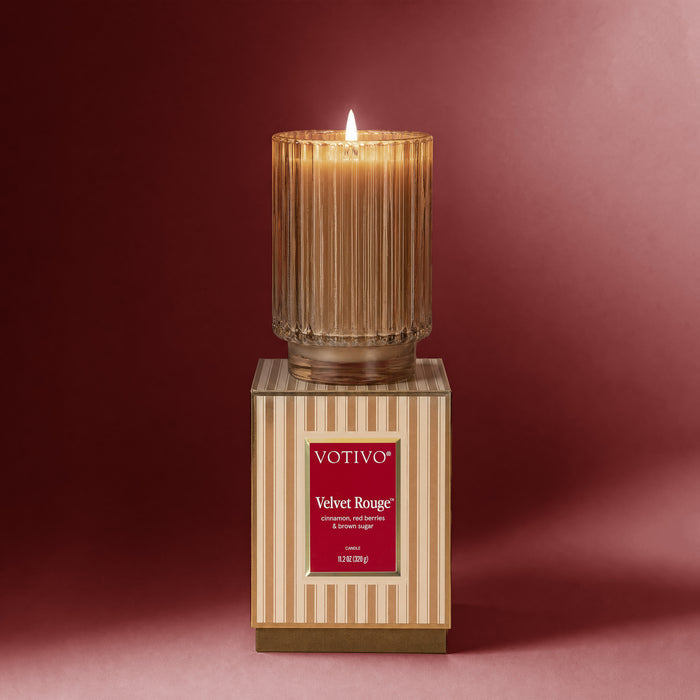 A lit Votivo Velvet Rouge Holiday Candle rests atop its coordinating striped box against a red backdrop. Housed in a ribbed glass jar and made from soy blend wax, the candle's label features fragrance notes of cardamom, red berries, and creamy woods.