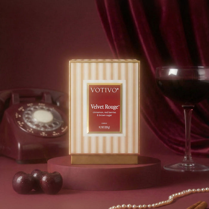 A Votivo Velvet Rouge Holiday Candle, made with a luxurious soy blend wax, is displayed on a platform next to a wine glass, vintage telephone, cherries, and a string of pearls against a rich burgundy velvet backdrop. The elegant arrangement creates a nostalgic atmosphere that exudes sophistication and luxury.