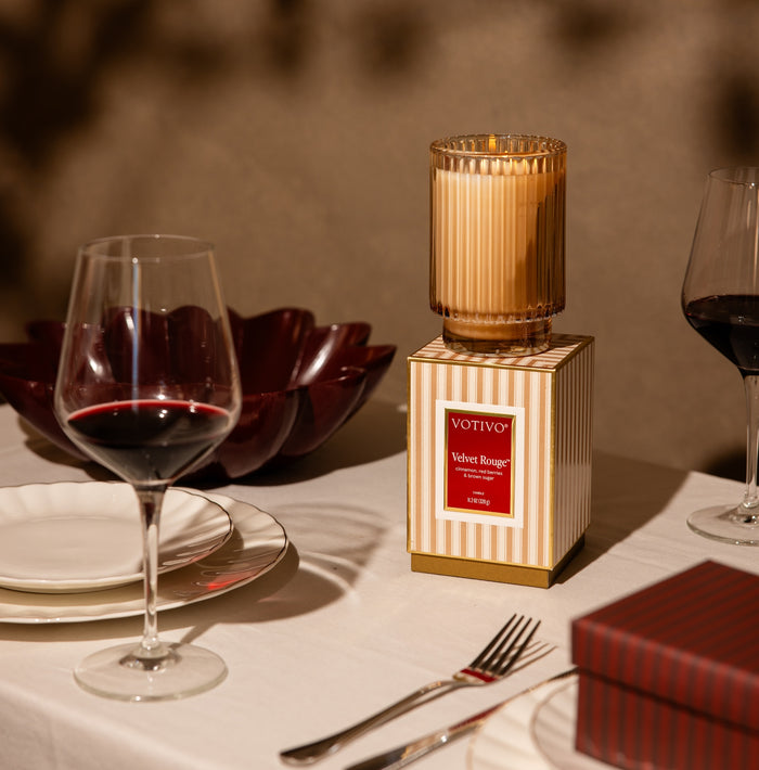A Votivo Velvet Rouge Holiday Candle sits atop its signature striped box on a set dining table with two glasses of red wine, white plates, a red bowl, and the coordinating box. Crafted from premium soy blend wax, the candle glows warmly as gentle lighting casts soft shadows across the cozy scene.