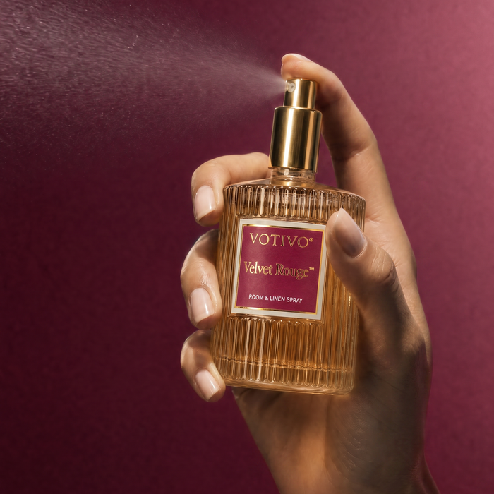 A hand spraying a Votivo Velvet Rouge room & linen spray, spraying a fine mist against a dark burgundy background.
