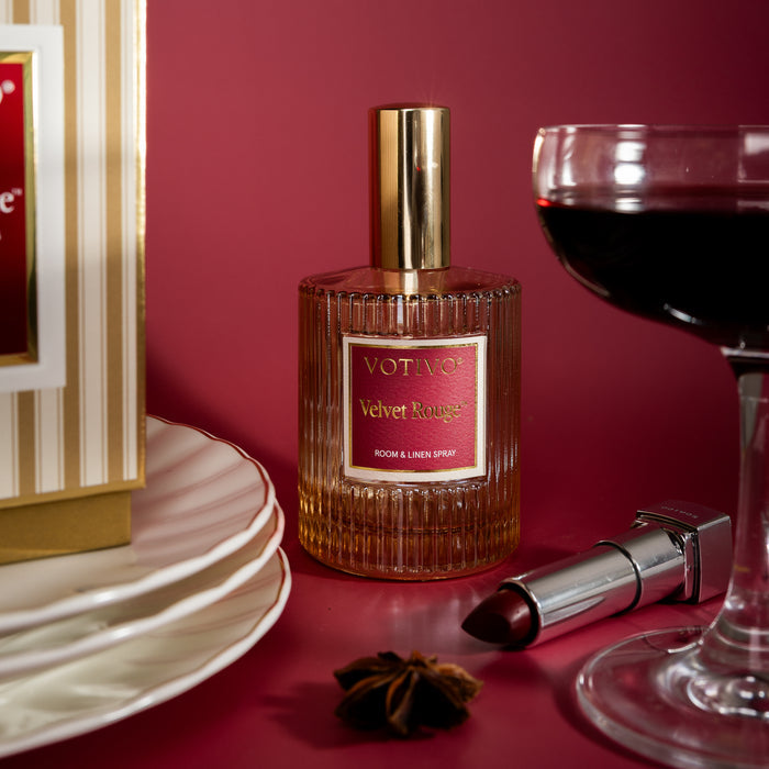 A bottle of Votivo Velvet Rouge Room & Linen Spray sits elegantly on a red surface beside a wine glass, stacked white plates, red lipstick, and a star anise.