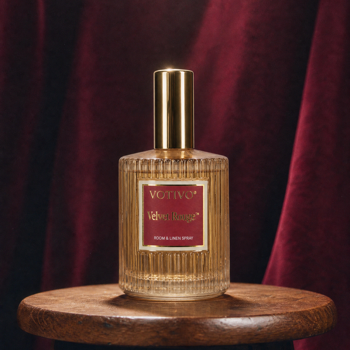 A Votivo Velvet Rouge Room & Linen Spray sitting on a wood stool against a burgundy background