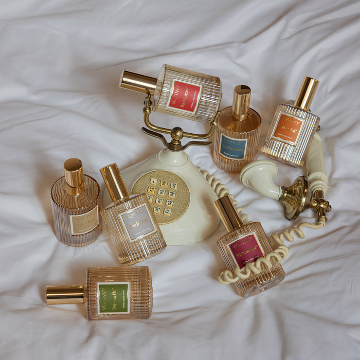Seven Votivo glass bottles, including Velvet Rouge Room & Linen Spray, with gold caps, colorful labels, and vertical ridges, are arranged around a vintage cream rotary phone on white wrinkled fabric.