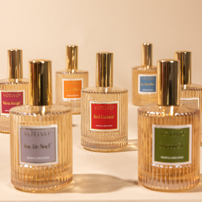 A selection of Votivo Velvet Rouge Room & Linen Spray bottles, each with unique colored labels and gold caps, are displayed on a beige surface, showcasing this elegant and fabric-safe fragrance.