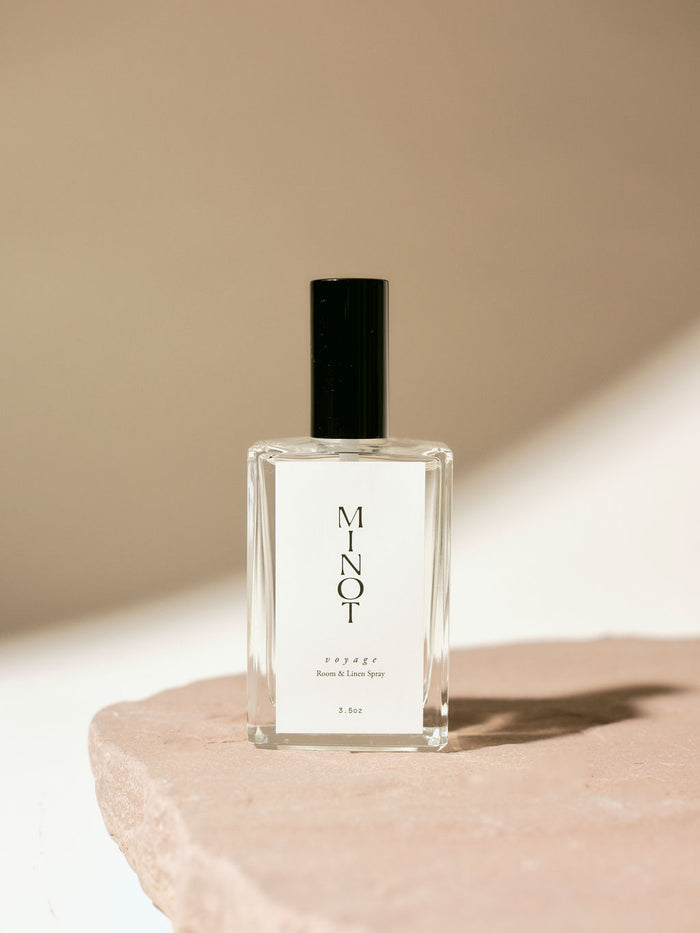 Clear glass bottle of MINOT Voyage room and linen spray with black cap, placed on a textured stone surface, highlighted by soft natural light.