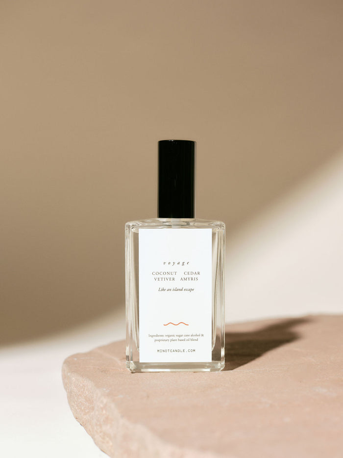 A bottle of MINOT's Voyage room and linen spray with coconut, cedar, vetiver, and amyris scents sits on a stone surface, with a minimalistic design and soft lighting casting gentle shadows.