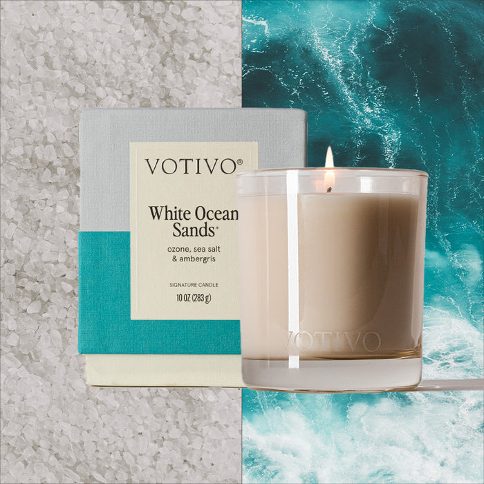 A Votivo White Ocean Sands 10 oz. Signature Candle and its box are side by side, set against white sea salt crystals and a turquoise ocean wave, with the lit soy blend wax candle in a clear glass jar.