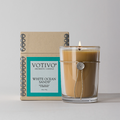 A Votivo White Ocean Sands 6.8 oz. Aromatic Candle with a burning wick sits before its matching aqua and beige box. Made from soy blend wax, its tied with twine and adorned with a round charm.