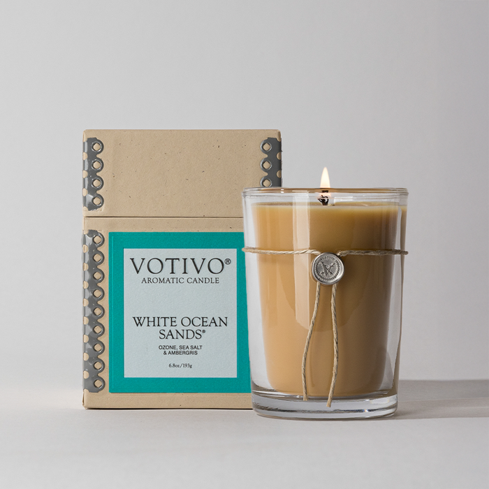 A Votivo White Ocean Sands 6.8 oz. Aromatic Candle with a burning wick sits before its matching aqua and beige box. Made from soy blend wax, its tied with twine and adorned with a round charm.