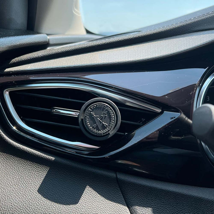 Close-up of a car air vent with a round black Votivo White Ocean Sands Car Fragrance (2 Pack) clipped on. The dashboard and part of the speedometer appear in sunlight, suggesting a refreshing scent inside.