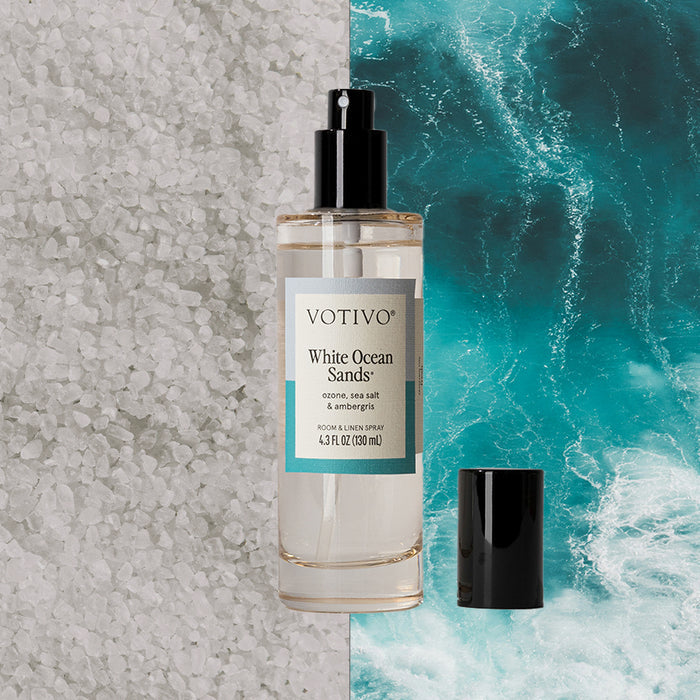 A Votivo White Ocean Sands Room & Linen Spray sits between coarse white salt and crashing blue waves, evoking a fresh scent. Its black cap rests nearby.