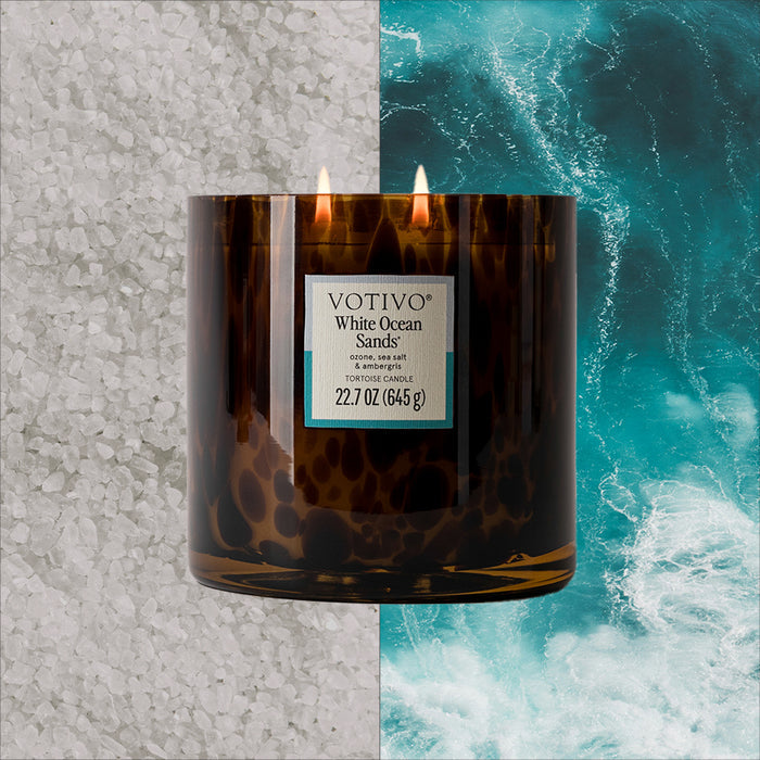 A brown glass Votivo White Ocean Sands Tortoise Candle is centered on a split background showing white sea salt on the left and ocean waves on the right.