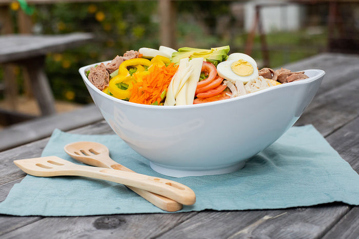 White bowl of colorful noodle salad with halved egg, sliced peppers, carrots and cucumber; wooden spoons on picnic table