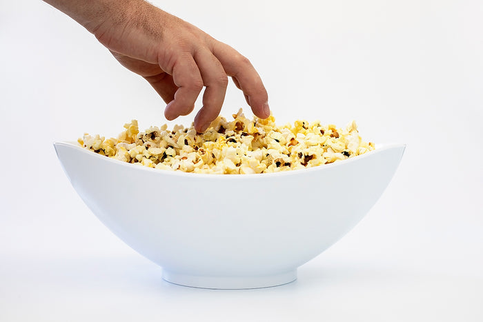 Side view of white oval salad bowl showing slanted asymmetrical rim, filled with popcorn with a hand reaching in, white background