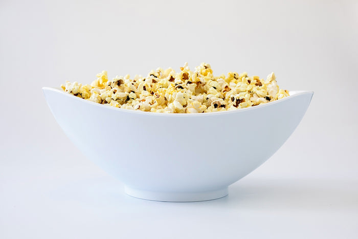 Side view of white oval salad bowl with slanted asymmetrical rim filled with buttered popcorn on white background