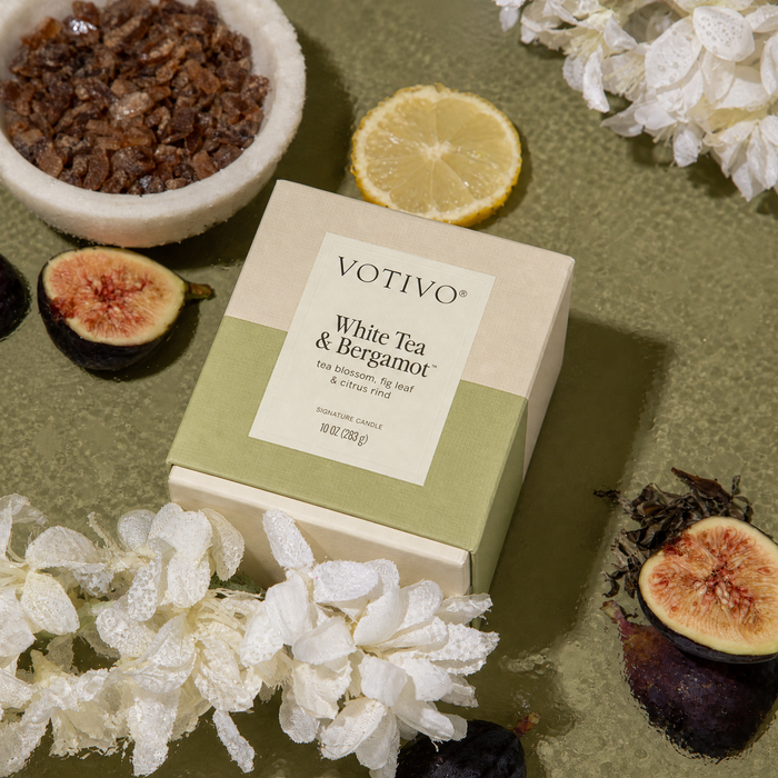 A Votivo White Tea & Bergamot 10 oz. Signature Candle box is surrounded by white flowers, a lemon slice, figs, tea leaves, and a bowl of brown sugar, all artfully arranged on a green textured surface. Crafted with soy blend wax by Votivo, this candle enhances the elegant display with its refined scent.