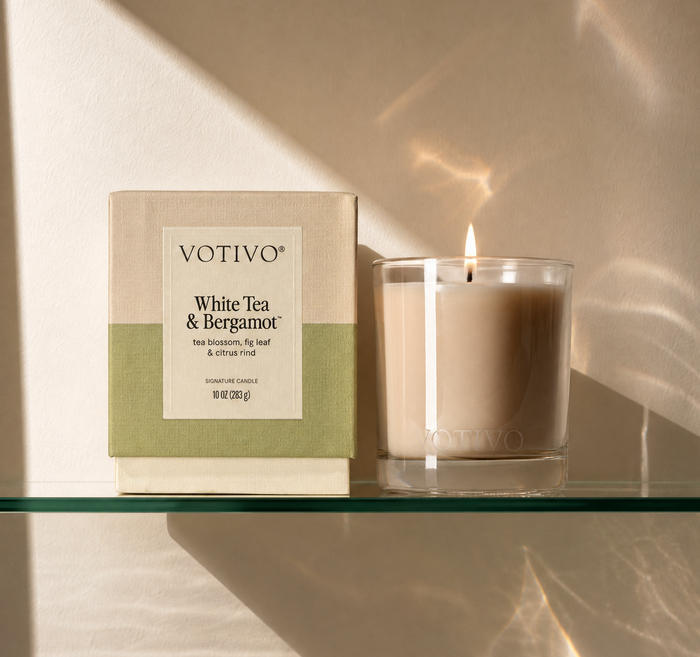 A Votivo White Tea & Bergamot 10 oz. Signature Candle, made from a premium soy blend wax, sits burning next to its box on a glass shelf as gentle sunlight reflects off the beige wall behind them.