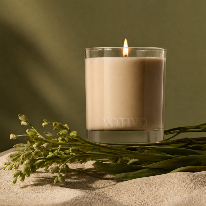 A lit White Tea & Bergamot 10 oz. Signature Candle by Votivo, made from a soy blend wax and housed in a clear glass jar, rests on a textured surface beside green sprigs, set against a muted green background.