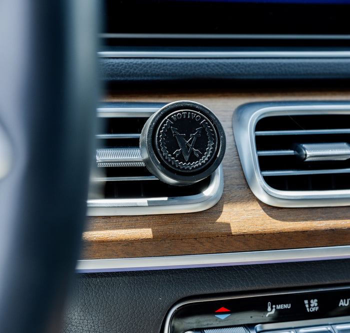 The Votivo White Tea & Bergamot Car Fragrance (2 Pack), featuring a black circular design with a butterfly logo, clips to your air vent to deliver fresh scent on-the-go. Wood-finish vent and dashboard buttons are visible in the image.