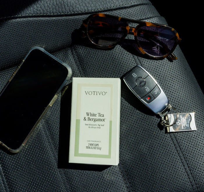 A box of Votivo White Tea & Bergamot Car Fragrance (2 Pack), a smartphone, tortoiseshell sunglasses, and a Mercedes-Benz key sit on a black leather seat—your essentials for fresh fragrance and style on the go.