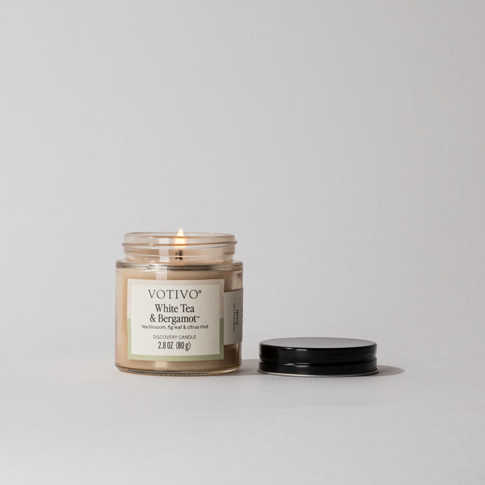 A lit Votivo White Tea & Bergamot Discovery Candle in a small glass jar sits beside its black lid on a neutral surface, showcasing the clean burn of its soy blend wax.