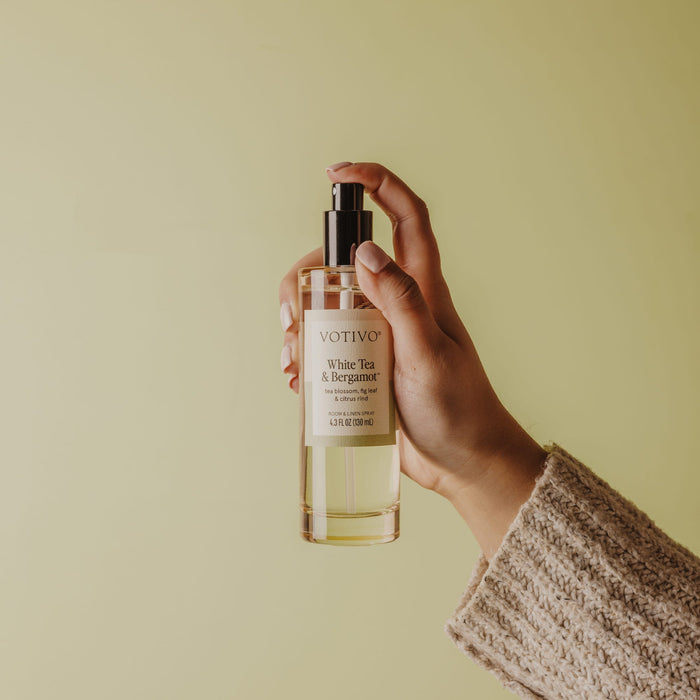 A hand in a beige knitted sleeve holds a clear Votivo White Tea & Bergamot Room & Linen Spray, fabric safe, against a light yellow-green background.