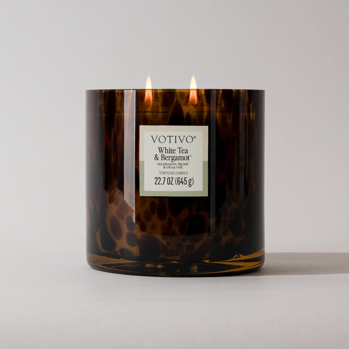 A brown, speckled tortoise jar holds a lit Votivo White Tea & Bergamot Tortoise Candle (22.7 oz), made from soy blend wax with two flames and a white front label displaying the weight.