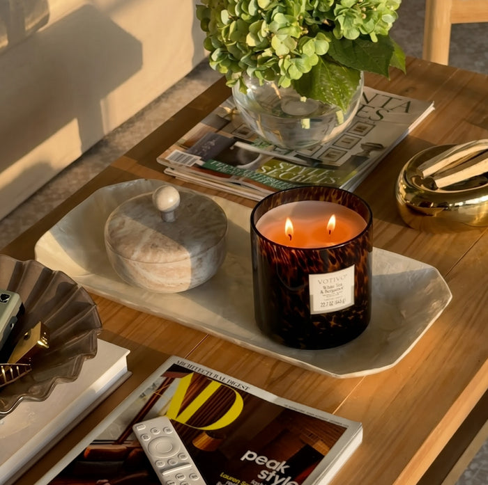A wooden coffee table displays the Votivo White Tea & Bergamot Tortoise Candle, crafted from a soy blend wax and lit beside a marble jar, magazines, a remote, a decorative leaf tray, and a vase of green hydrangeas—all arranged neatly in warm sunlight.