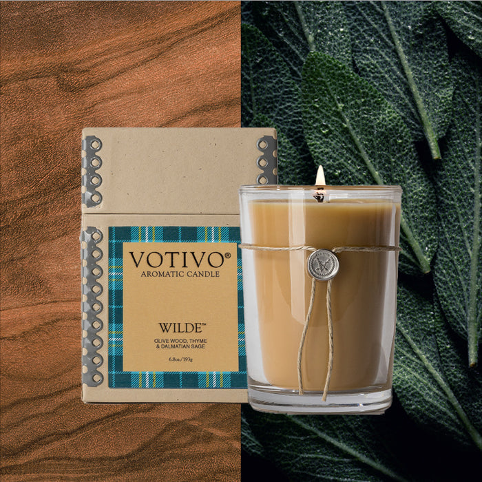 The Votivo Wilde 6.8 oz. Aromatic Candle features a unique scent and comes in a glass jar wrapped with twine, shown next to its box on brown wood grain with green sage leaves for an inviting look.