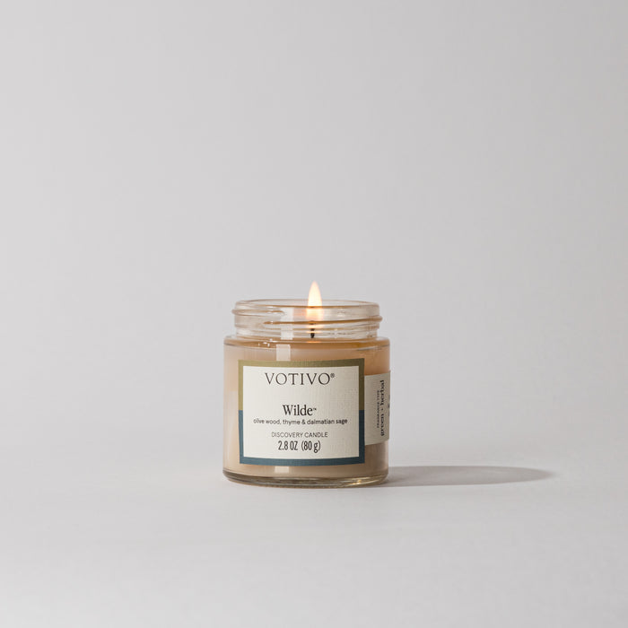 A Votivo Wilde Discovery Candle burns softly in a clear glass jar, filling the air with lasting fragrance against a plain white background.