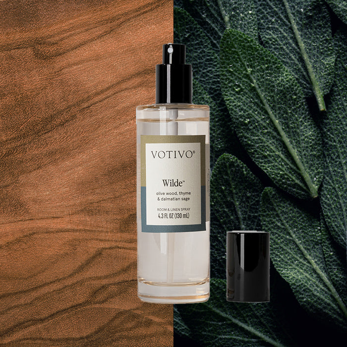 A clear bottle of Votivo Wilde Room & Linen Spray with a black cap is set against rich wood grain and green sage leaves, reflecting its signature rosemary sage fragrance.